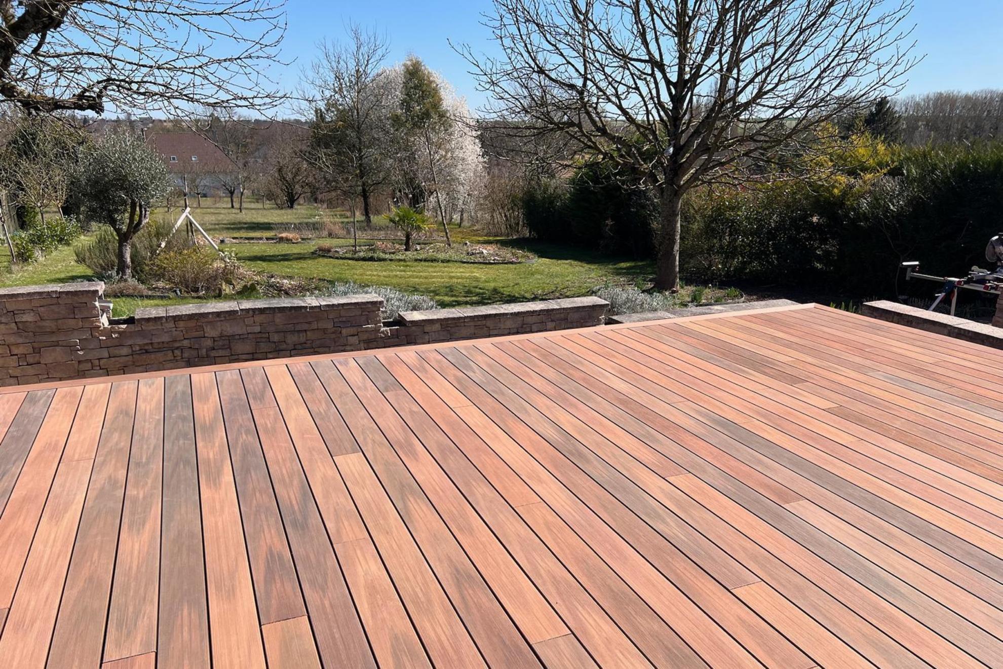 Terrasse en bois composite Silvadec installée dans un jardin de campagne avec muret en pierre et vue sur un verger, aménagement paysager par Agave Paysage