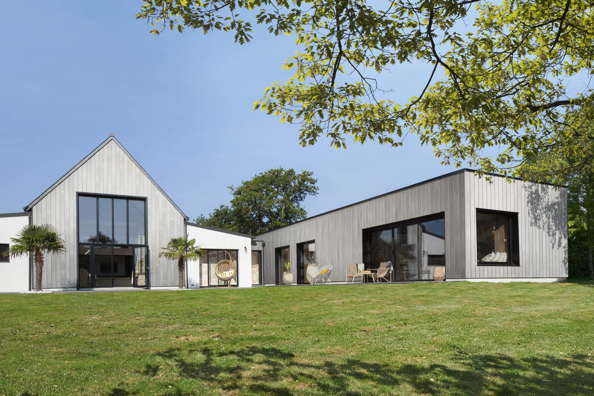 Zeitgenössisches Haus mit Fassadenverkleidung aus Holzverbundwerkstoff Silvadec, bestehend aus zwei separaten Volumen mit Giebeldach und Flachdach, umgeben von einem grünen Garten