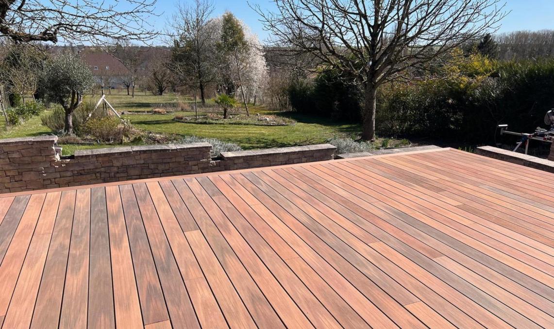 Terrasse en bois composite Silvadec installée dans un jardin de campagne avec muret en pierre et vue sur un verger, aménagement paysager par Agave Paysage